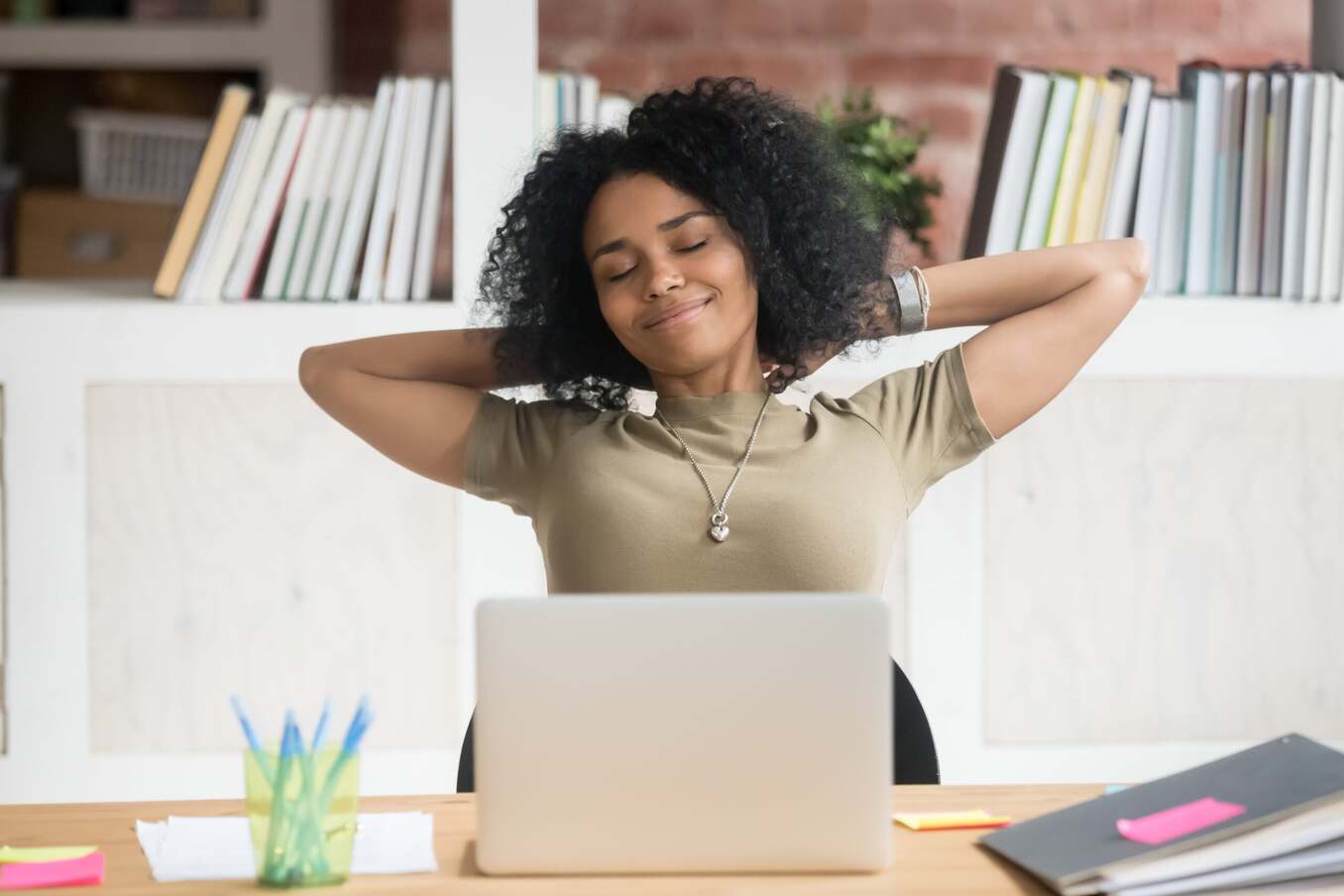 A woman sitting at a desk with a laptop, smiling with her eyes closed and hands behind her head as she stretches and takes a break