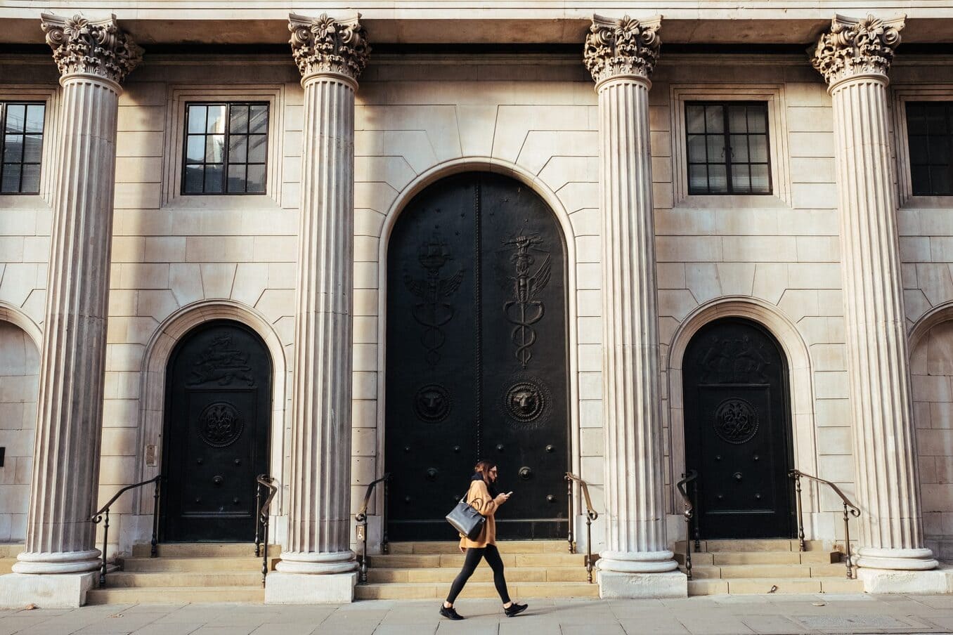 A woman walking along a sidewalk past a large stone building with tall Corinthian columns and ornate black arched doors