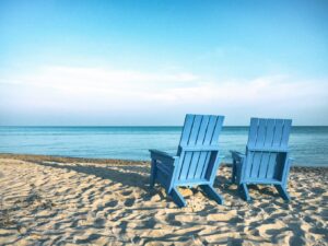 Two empty light blue Adirondack chairs sitting on a sandy beach, facing a calm, turquoise ocean under a clear blue sky. The scene is peaceful and sunny, with soft shadows on the sand.