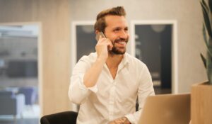 A smiling man with a beard and a white shirt sitting at a desk, talking on a smartphone in a modern office environment