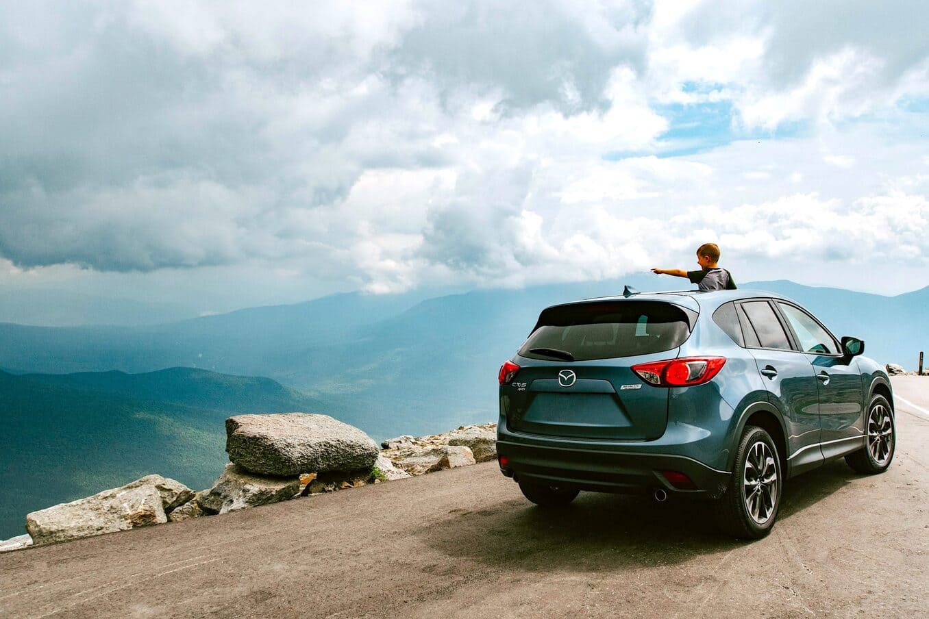 A young boy looking out from the sunroof of a blue SUV parked at a mountain overlook, pointing toward a vast valley under a cloudy sky