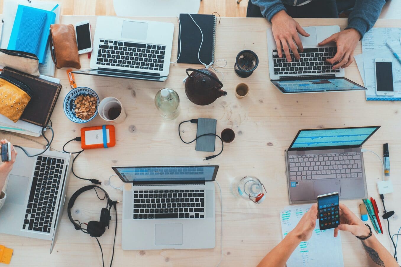 An overhead view of a busy wooden communal table where several people are working together. The desk is cluttered with multiple laptops showing data charts, smartphones, headphones, notebooks, and coffee cups, capturing a high-energy collaborative workspace