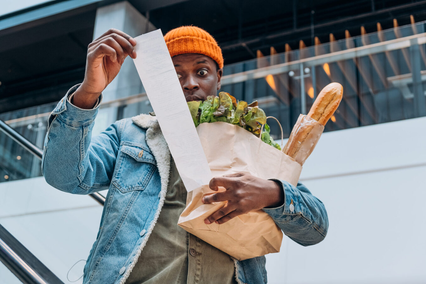 A man with a surprised expression holding a brown paper bag of groceries and looking at a very long receipt