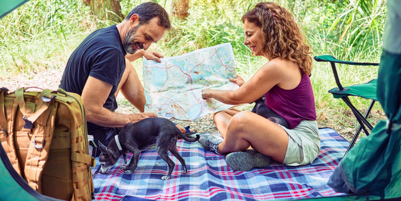 A middle-aged man and woman sitting on a blue plaid blanket outside their tent while camping in a forest. They are smiling and looking at a large paper map together, while a small black dog explores the blanket near their hiking gear and backpacks.