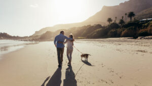 A view from behind of a middle-aged couple walking arm-in-arm along a wide, sandy beach at sunset. They are walking a beagle on a leash toward a backdrop of lush green hills and palm trees, with the warm sun creating long shadows on the sand.