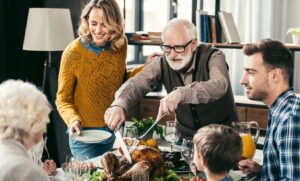 A senior man with glasses and a white beard carving a roasted turkey at a crowded dinner table. His family, including a young woman in a yellow sweater holding a plate and a man in a plaid shirt, watch with smiles. The scene is a warm, indoor family gathering with wine glasses and side dishes visible.