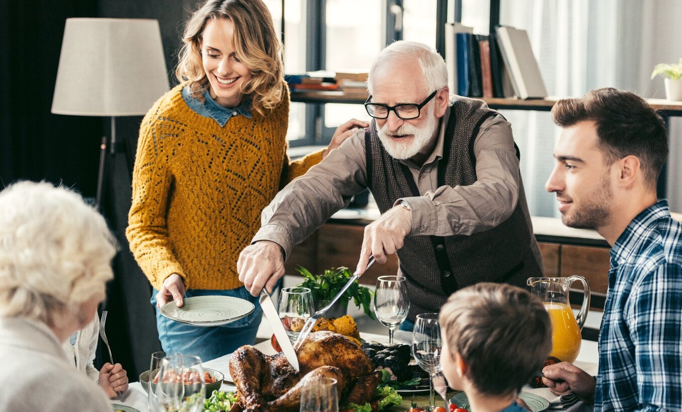 A senior man with glasses and a white beard carving a roasted turkey at a crowded dinner table. His family, including a young woman in a yellow sweater holding a plate and a man in a plaid shirt, watch with smiles. The scene is a warm, indoor family gathering with wine glasses and side dishes visible.
