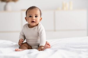 An adorable baby sitting on a white bed in a bright, modern room.