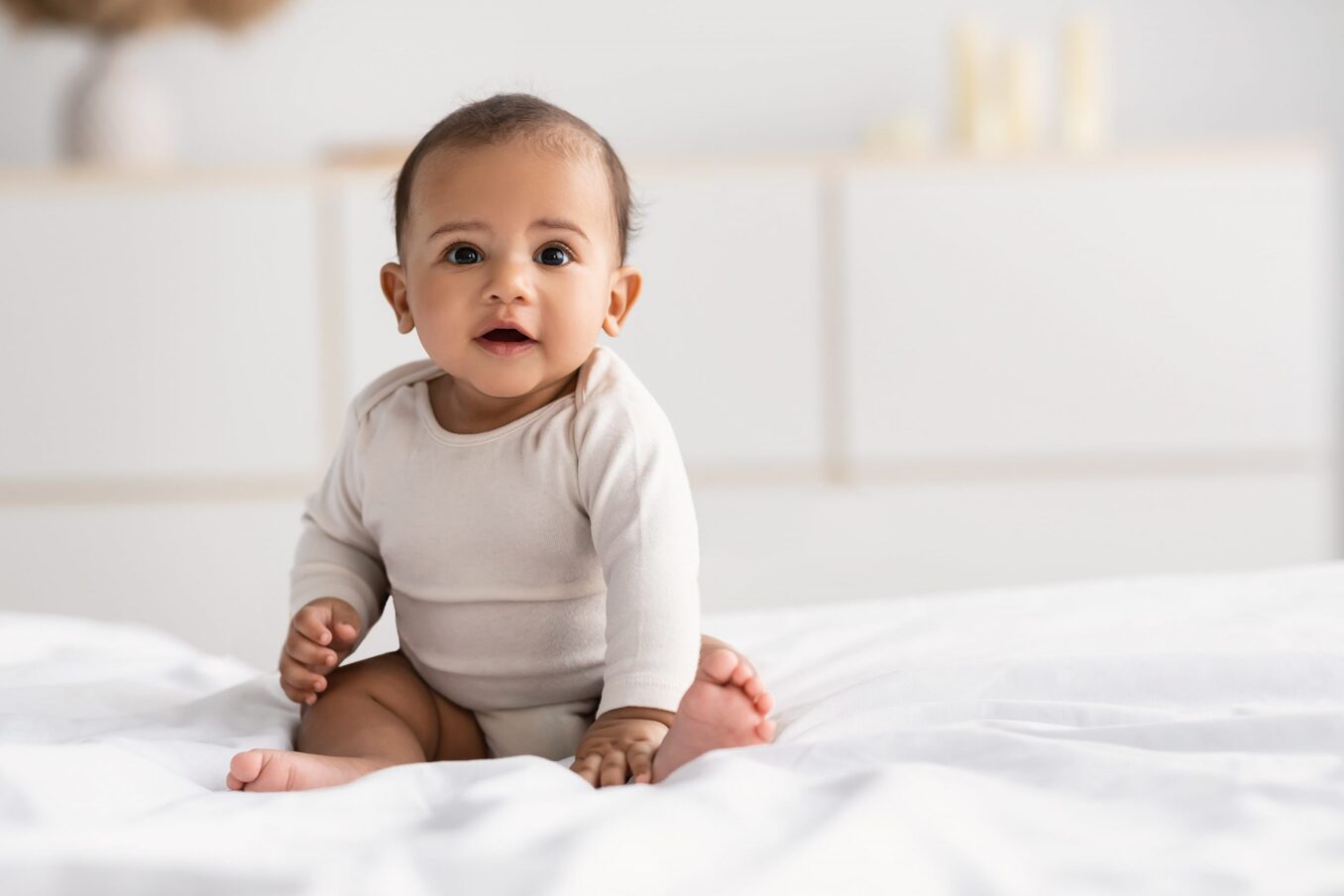An adorable baby sitting on a white bed in a bright, modern room.