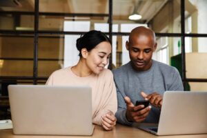 Couple in front of their laptops on a table. The man is holding a phone and showing something to his partner.