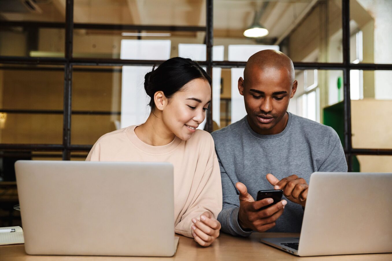 Couple in front of their laptops on a table. The man is holding a phone and showing something to his partner.
