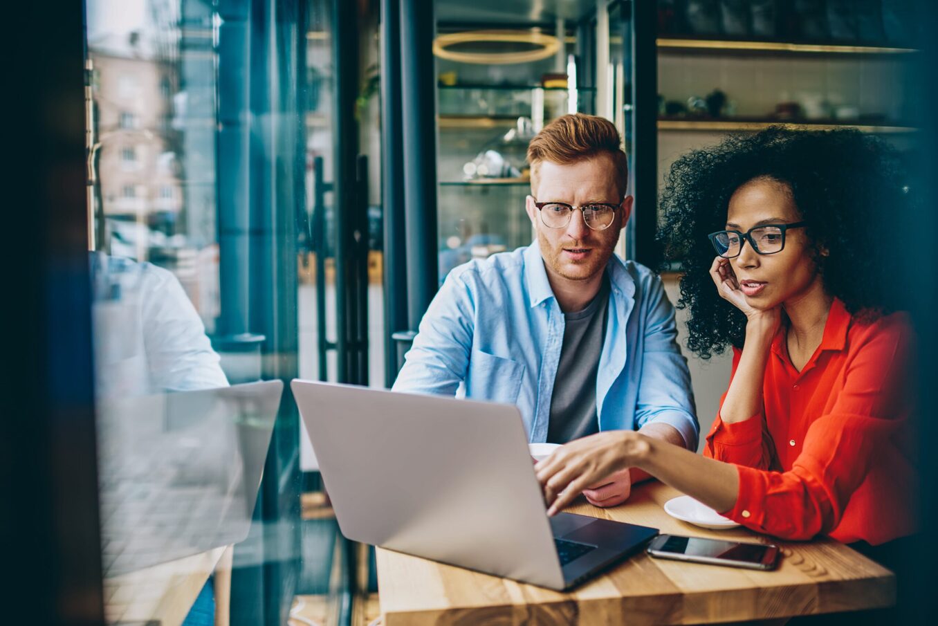 A diverse man and woman sitting at a small wooden table in a cafe, looking intently at a laptop screen together. The man has red hair and glasses, wearing a light blue shirt, while the woman has dark curly hair and glasses, wearing a vibrant orange blouse and pointing at the screen.