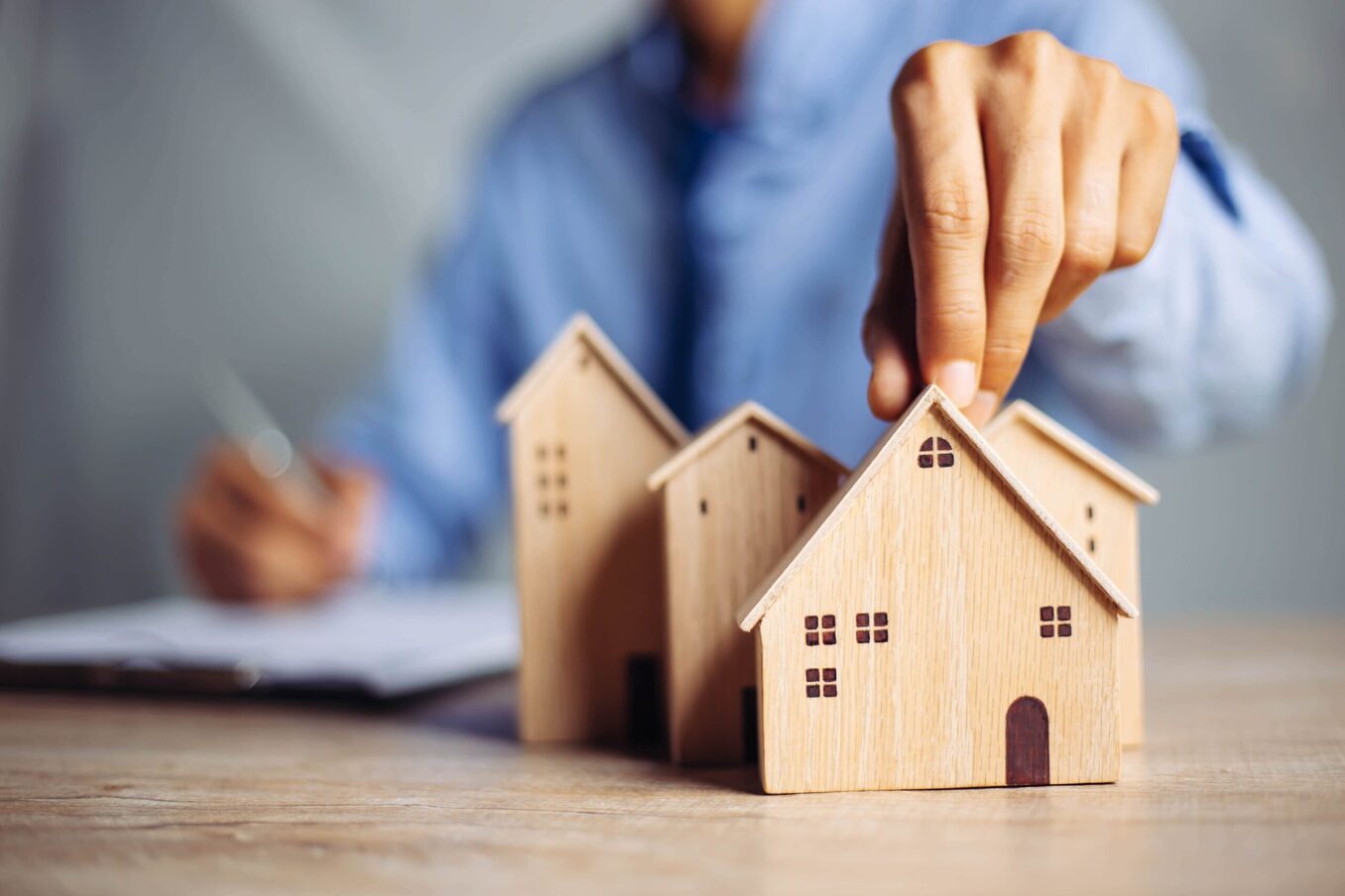 A close-up of a person’s hand placing a small wooden house model among a cluster of other wooden houses on a desk. In the blurred background, a person in a blue shirt is writing on a clipboard, symbolizing real estate planning, home buying, or property investment.