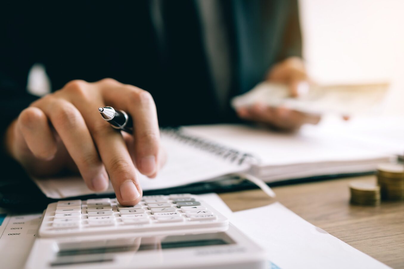 A close-up of a person's hands using a white calculator while holding a pen over an open notebook. In the blurred background, they are holding a fan of cash next to stacks of coins on a wooden desk, symbolizing accounting, budgeting, or financial management.