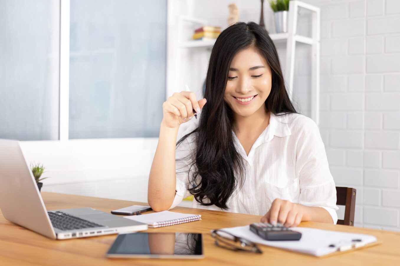 A woman smiling while using a calculator and laptop at an office desk.