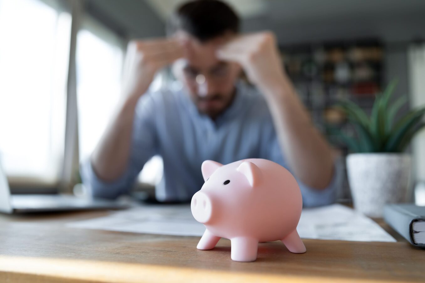 Stressed young male professional holding his head while looking at documents. Piggy bank in front