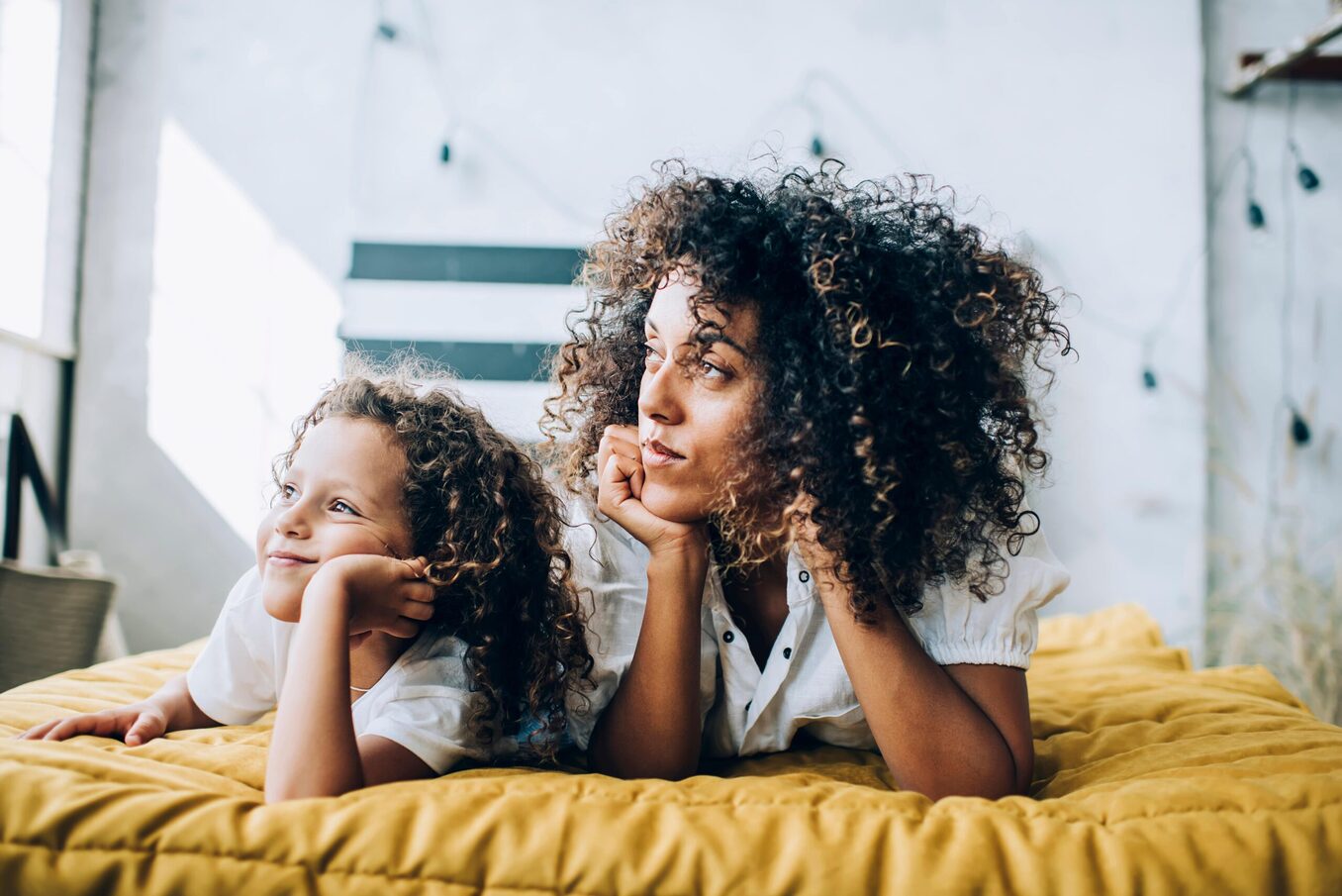A woman and a young girl, both with voluminous curly hair, lying on their stomachs on a yellow textured bedspread. They are both resting their chins on their hands and looking off-camera with thoughtful, happy expressions in a bright, modern room.