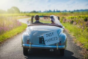 A rearview shot of a newlywed couple driving a vintage light blue convertible down a winding country road. A "Just Married" sign is attached to the back of the car, and the bride's white veil flutters in the wind as they drive through a sunny landscape.