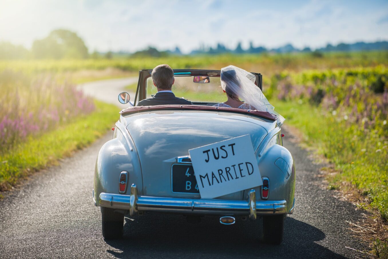 A rearview shot of a newlywed couple driving a vintage light blue convertible down a winding country road. A "Just Married" sign is attached to the back of the car, and the bride's white veil flutters in the wind as they drive through a sunny landscape.