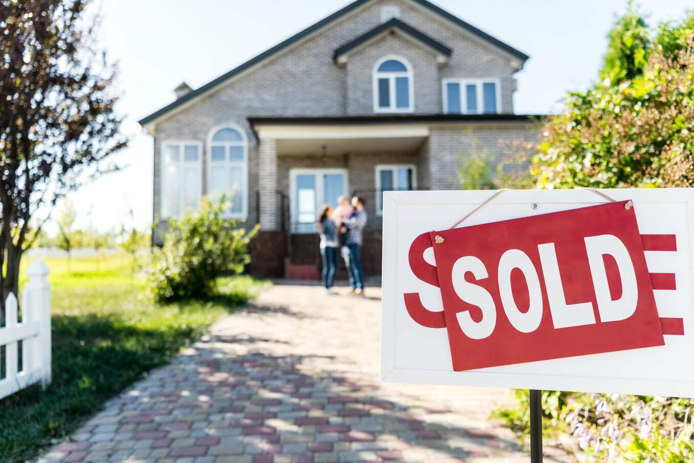 A close-up of a white real estate sign with a red "SOLD" placard in the foreground. In the blurred background, a happy family of three stands in front of a modern two-story brick house, celebrating their new home purchase on a sunny day.