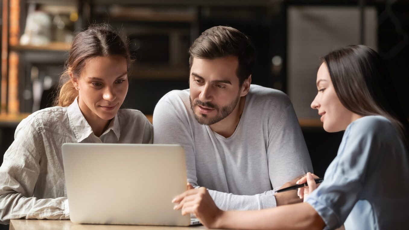 A professional woman in a light blue shirt points to a laptop screen while explaining information to a young couple. The man and woman lean in with focused expressions as they review the details together at a table in a modern office or cafe setting.