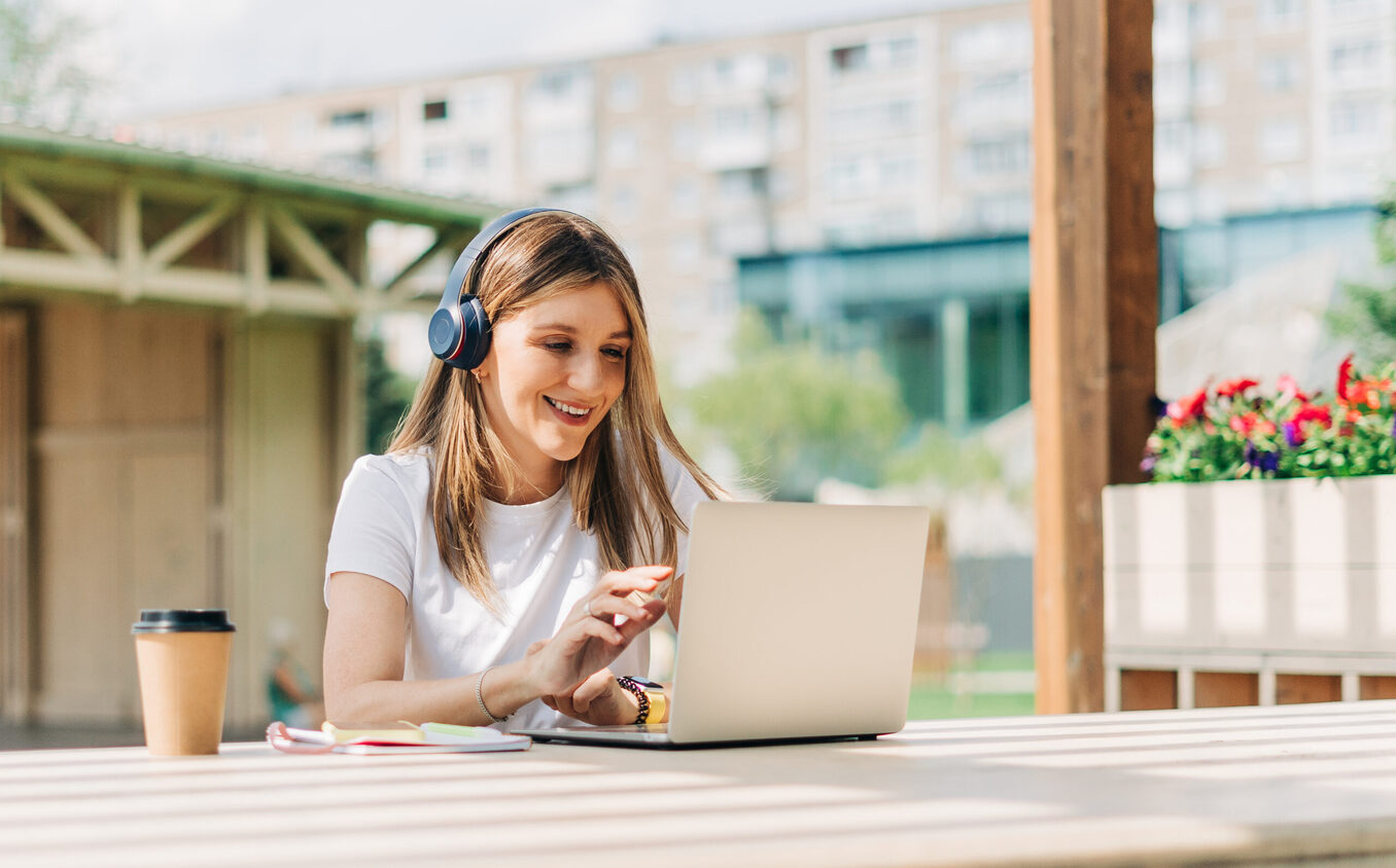 Young hipster Woman in headphones using laptop, having a video call chat outside in park. Happy and smiling typing at notebook with coffee cup.