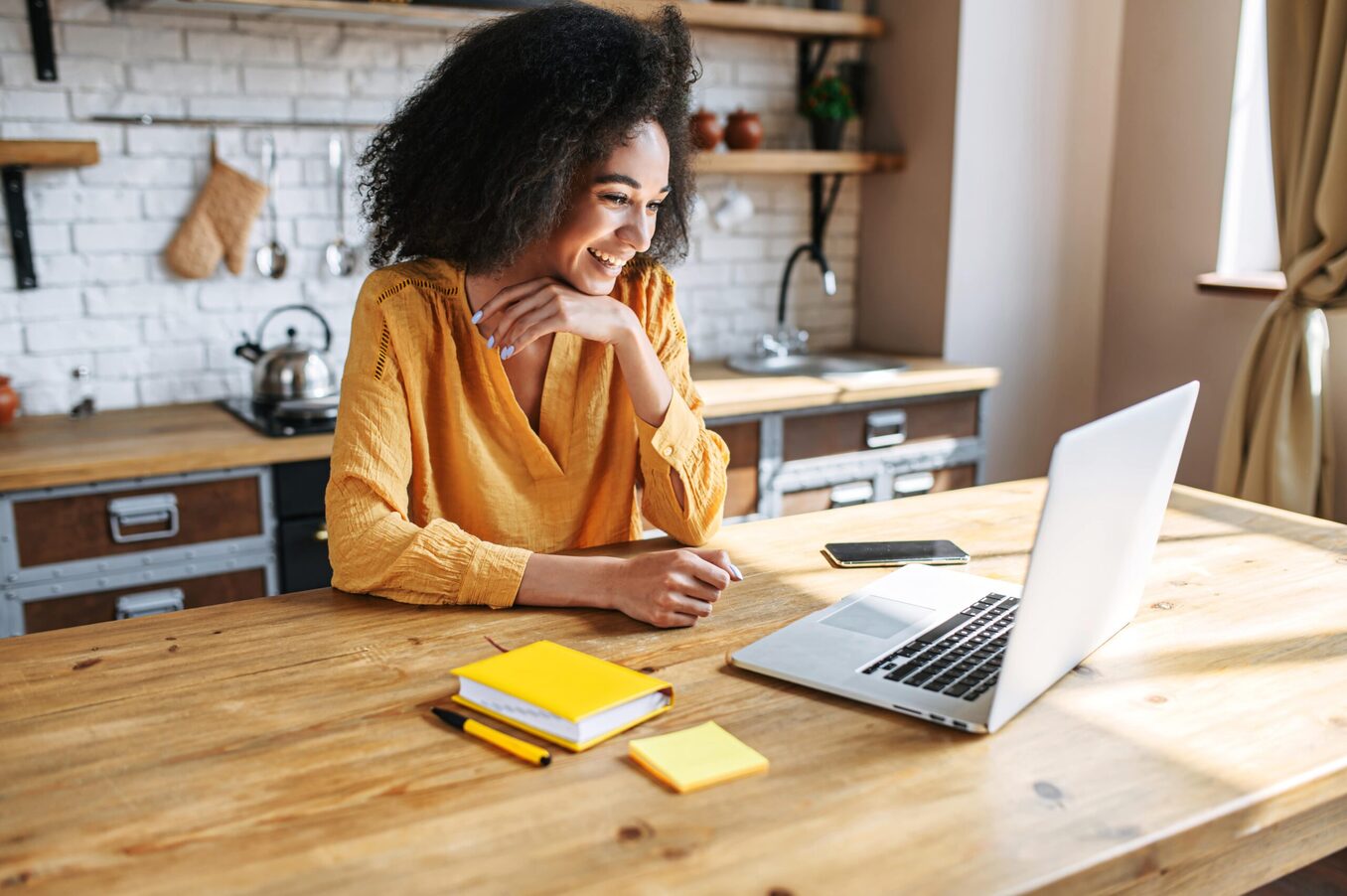 A smiling young woman with an afro hairstyle sitting at a light wooden kitchen table, looking at her laptop. She is wearing a mustard-yellow blouse and has a yellow notebook, pen, and smartphone on the table in front of her. The background shows a modern, rustic-style kitchen with white brick walls.