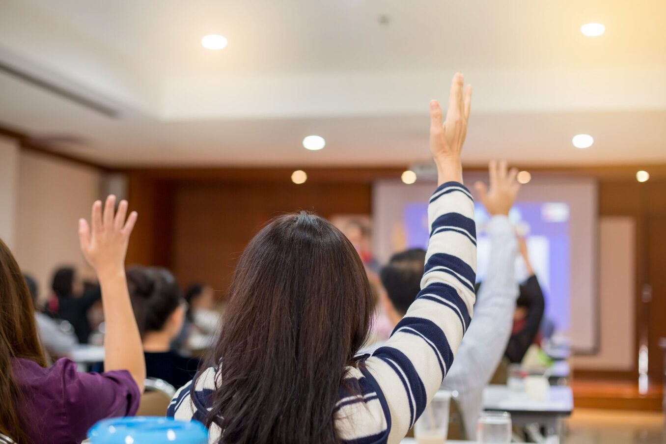 Room full of people raising hands during a lecture or presentation