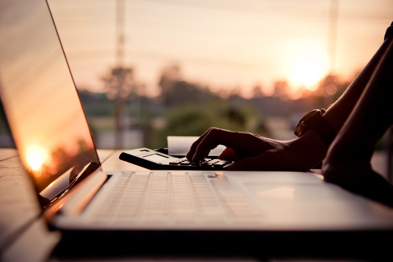 Person using their laptop and calculator on a table outside around sunset.