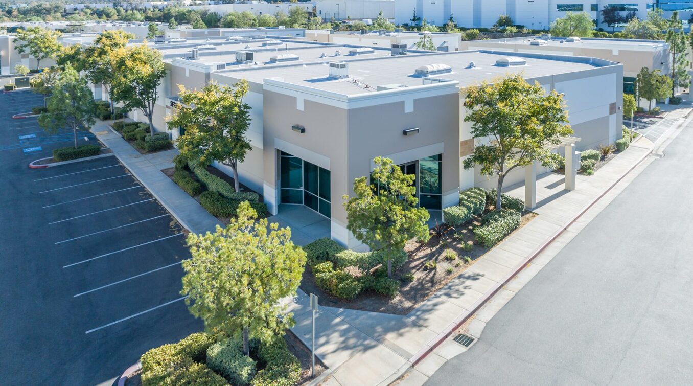 An aerial high-angle view of a modern one-story commercial office building in a suburban business park. The building features beige and white exterior walls with large glass windows, surrounded by paved parking lots, green trees, and manicured landscaping under a clear sky.