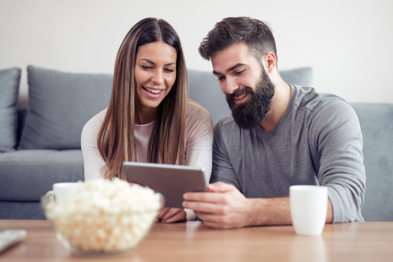 A happy couple sitting at home and watching content on a tablet with popcorn.