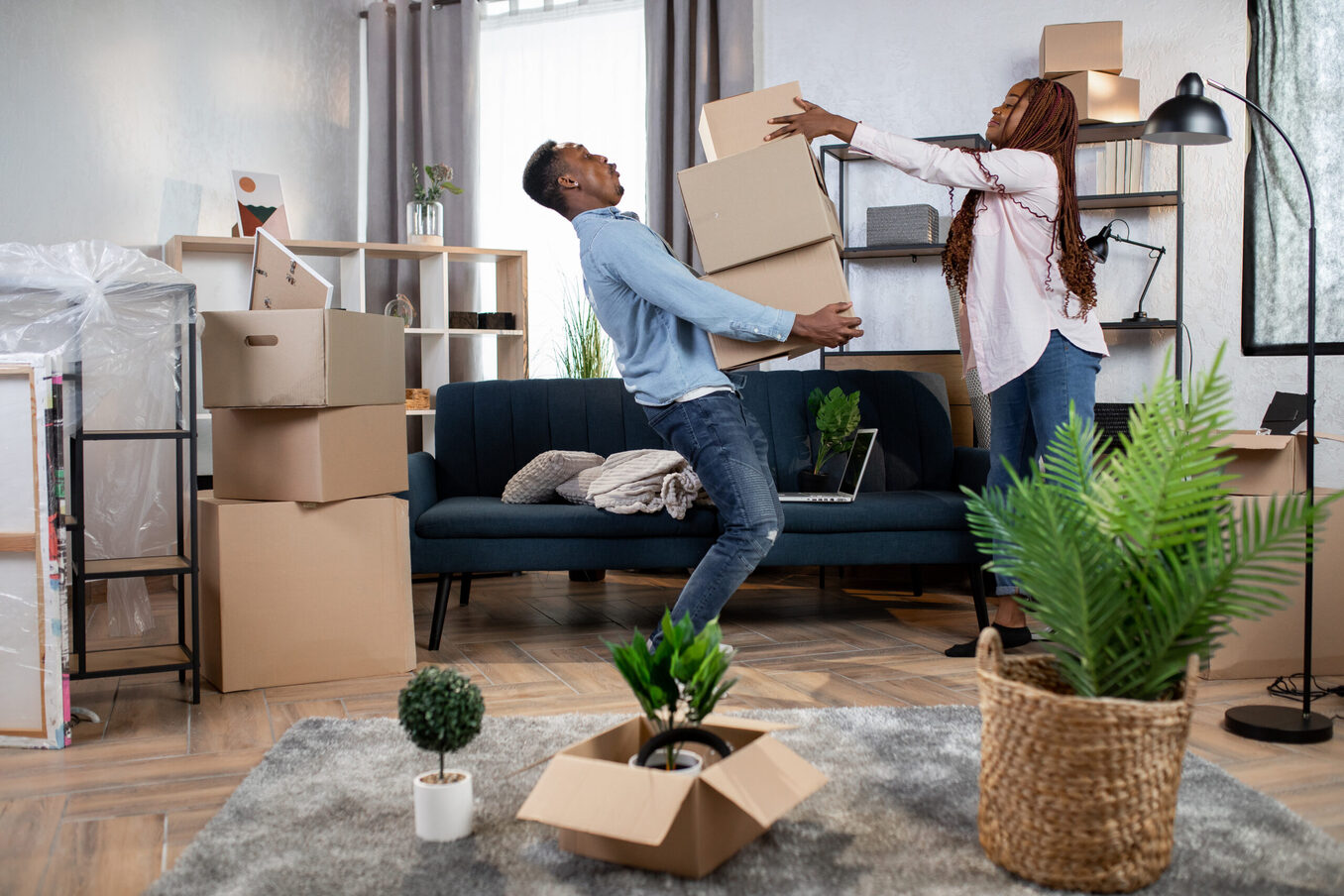 A young couple playfully struggling with a stack of heavy boxes while moving into a new home