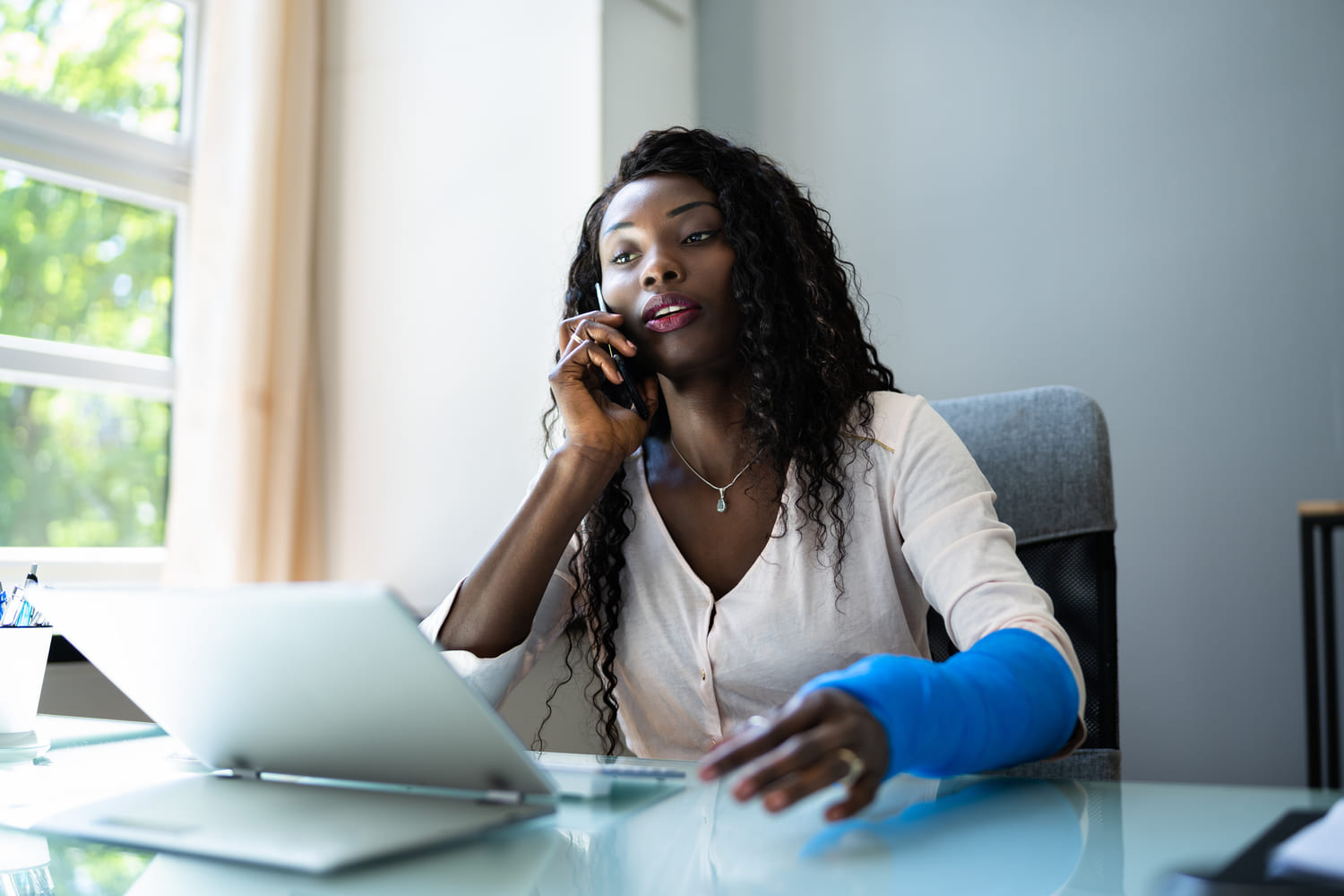 An office worker with her arm in a blue cast talking on the phone at her desk.