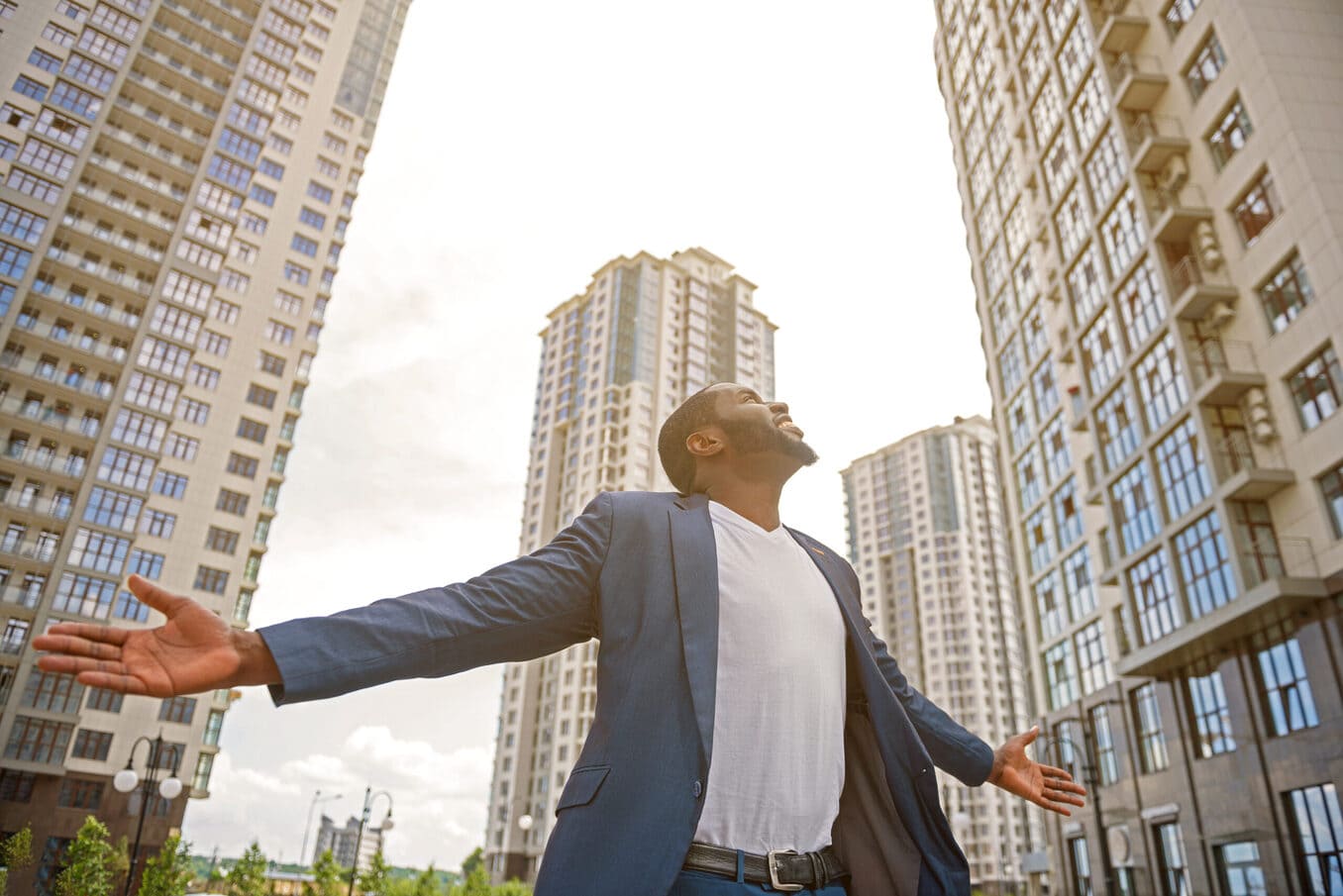 A man in a blue blazer standing with his arms outstretched and head tilted back, looking up toward tall apartment buildings under a bright sky
