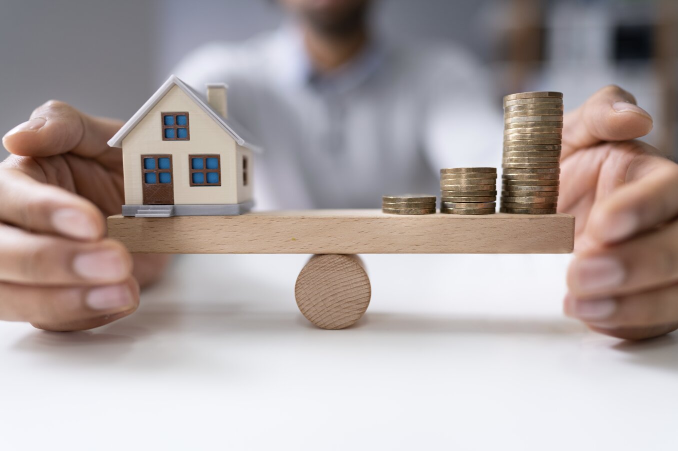 Businessperson's Hand Protecting Balance Between House Model And Increasing Stacked Coins On Seesaw