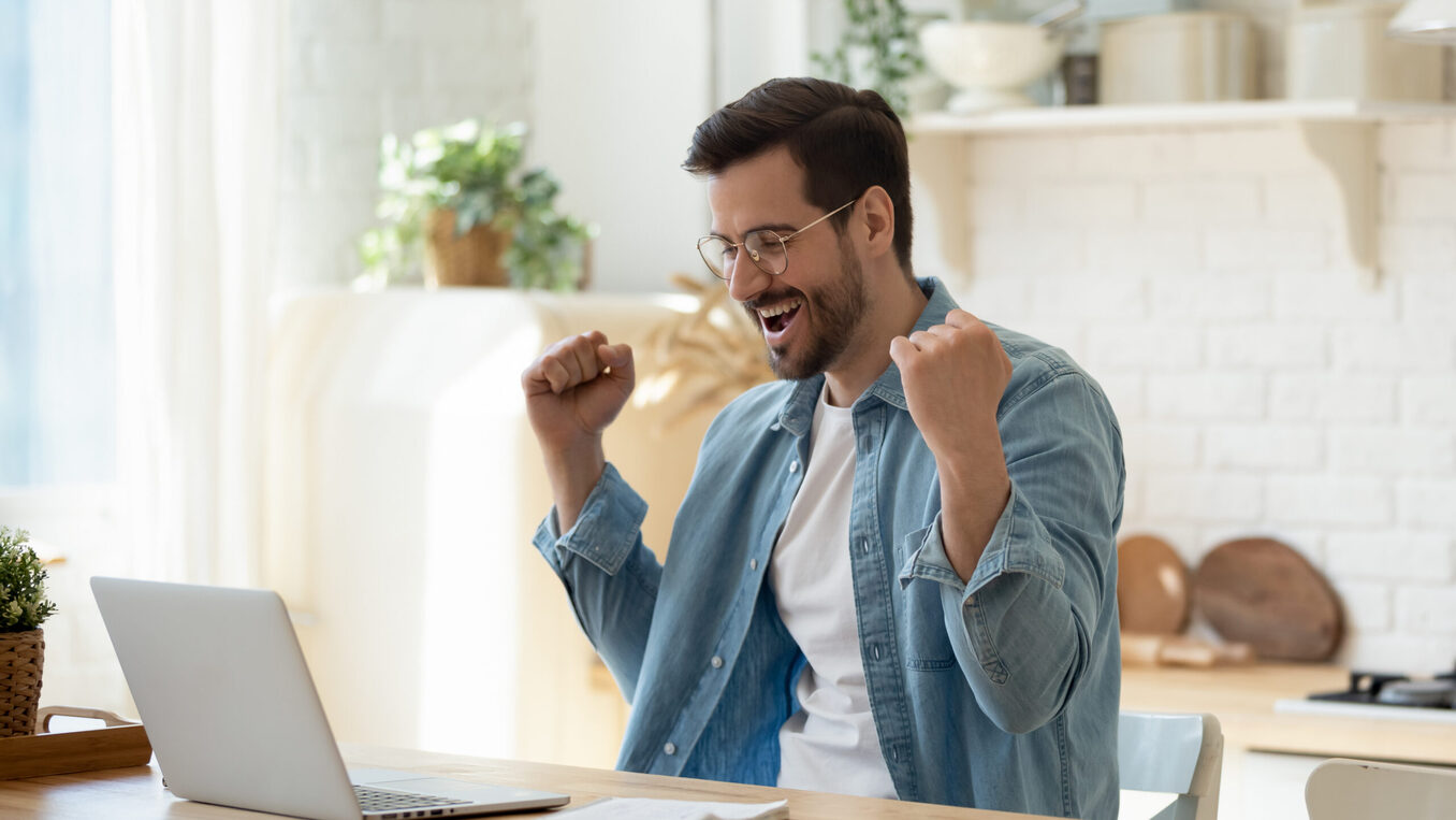 Happy guy celebrate great news opportunity got read by e-mail. Excited man sit in kitchen look at laptop screen with clenched fists feels incredible amazed received good notice. Sale, discount concept