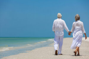 Rear view of happy senior man and woman couple walking and holding hands on a deserted tropical beach with bright clear blue sky