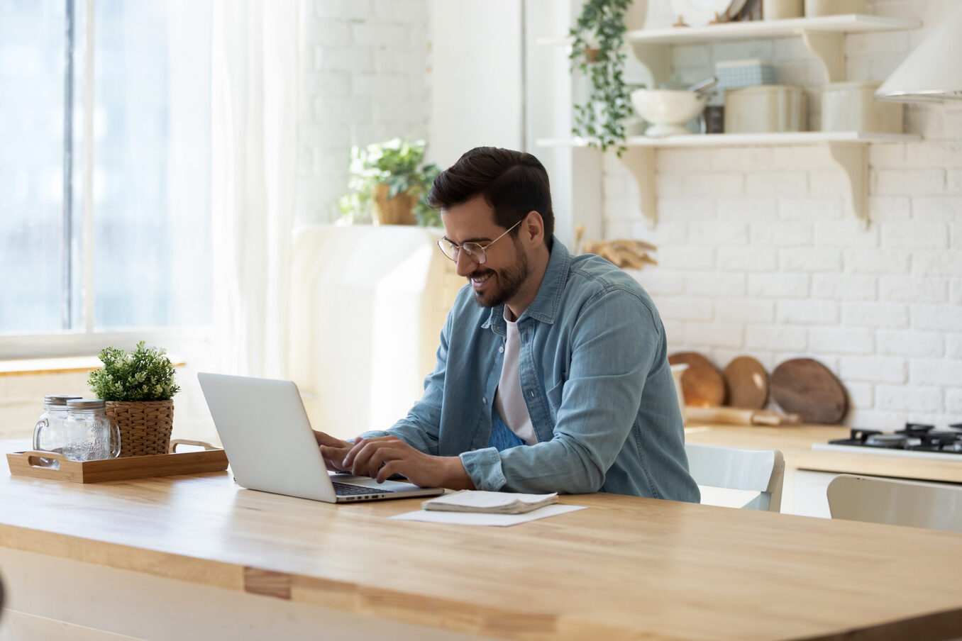 Focused millennial Caucasian man sit at table at home kitchen look at laptop screen working distant. Young male use computer, texting or messaging online on device. Communication, technology concept.