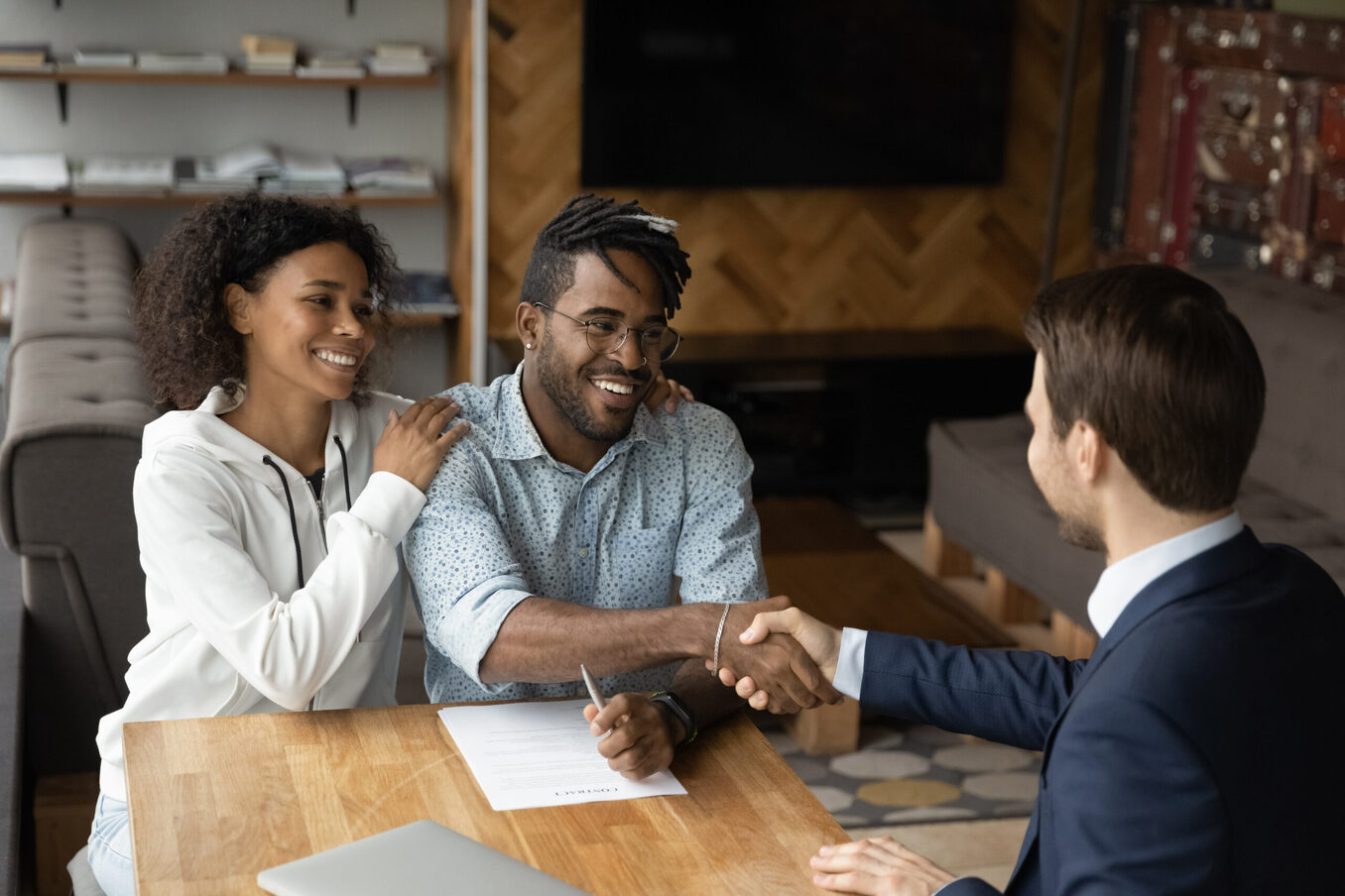 A smiling couple sitting at a wooden table, with the man shaking hands with a professional advisor across from them after signing a document