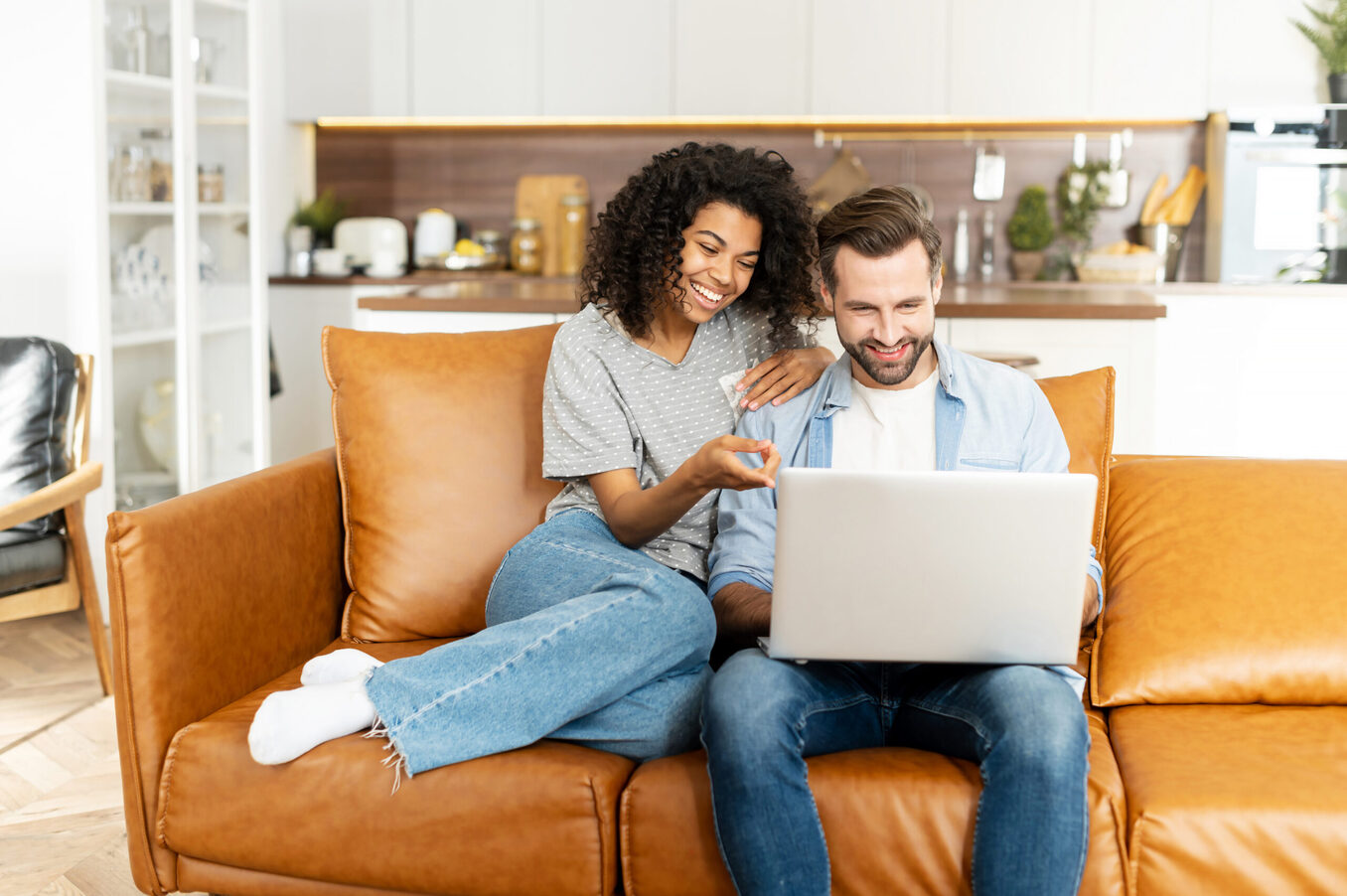 Cheerful diverse couple in love spends leisure time online with a laptop at home. Young woman and man look at the laptop screen and laugh sitting on the sofa, watching comedy movies, funny videos