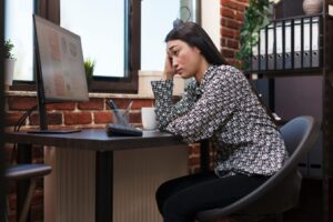 A woman sitting at a desk in a home office, looking stressed and resting her head on her hand while staring at financial charts on a computer monitor