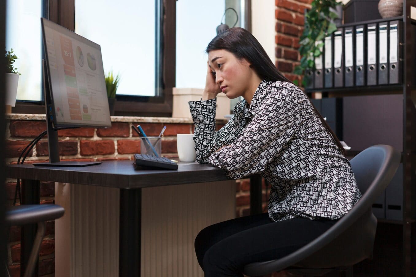 A woman sitting at a desk in a home office, looking stressed and resting her head on her hand while staring at financial charts on a computer monitor