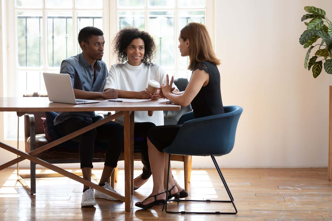 Young African American husband and wife sit at desk talk with female real estate agent discuss buying house together, biracial couple meet with designer architect or broker consult in modern office