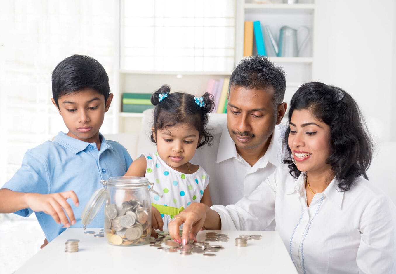 A family of four sitting together at a table, with two young children and their parents counting coins and putting them into a glass savings jar