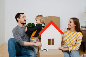 Family moved into a new apartment, parents looking at their son. They surrounded by multiple sealed cardboard boxes. Mother holding toy paper house in hands, symbol of a new home.
