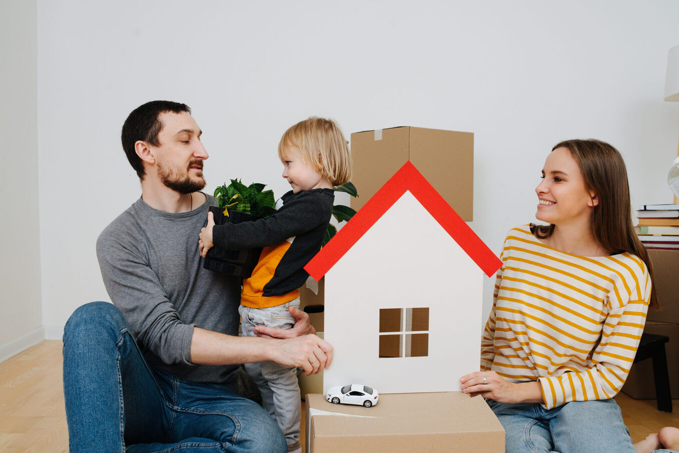 Family moved into a new apartment, parents looking at their son. They surrounded by multiple sealed cardboard boxes. Mother holding toy paper house in hands, symbol of a new home.