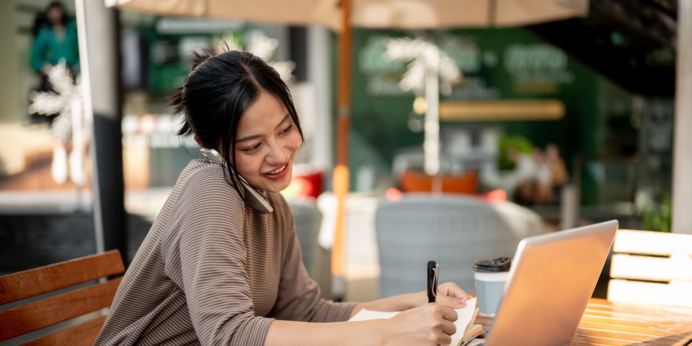 A smiling woman talking on her smartphone while taking notes in a notebook and working on a laptop at an outdoor cafe table.