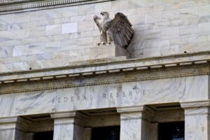 A low-angle shot of the white marble exterior of the Federal Reserve building, featuring the engraved "FEDERAL RESERVE" title and a large stone American eagle sculpture with outstretched wings perched above the entrance.