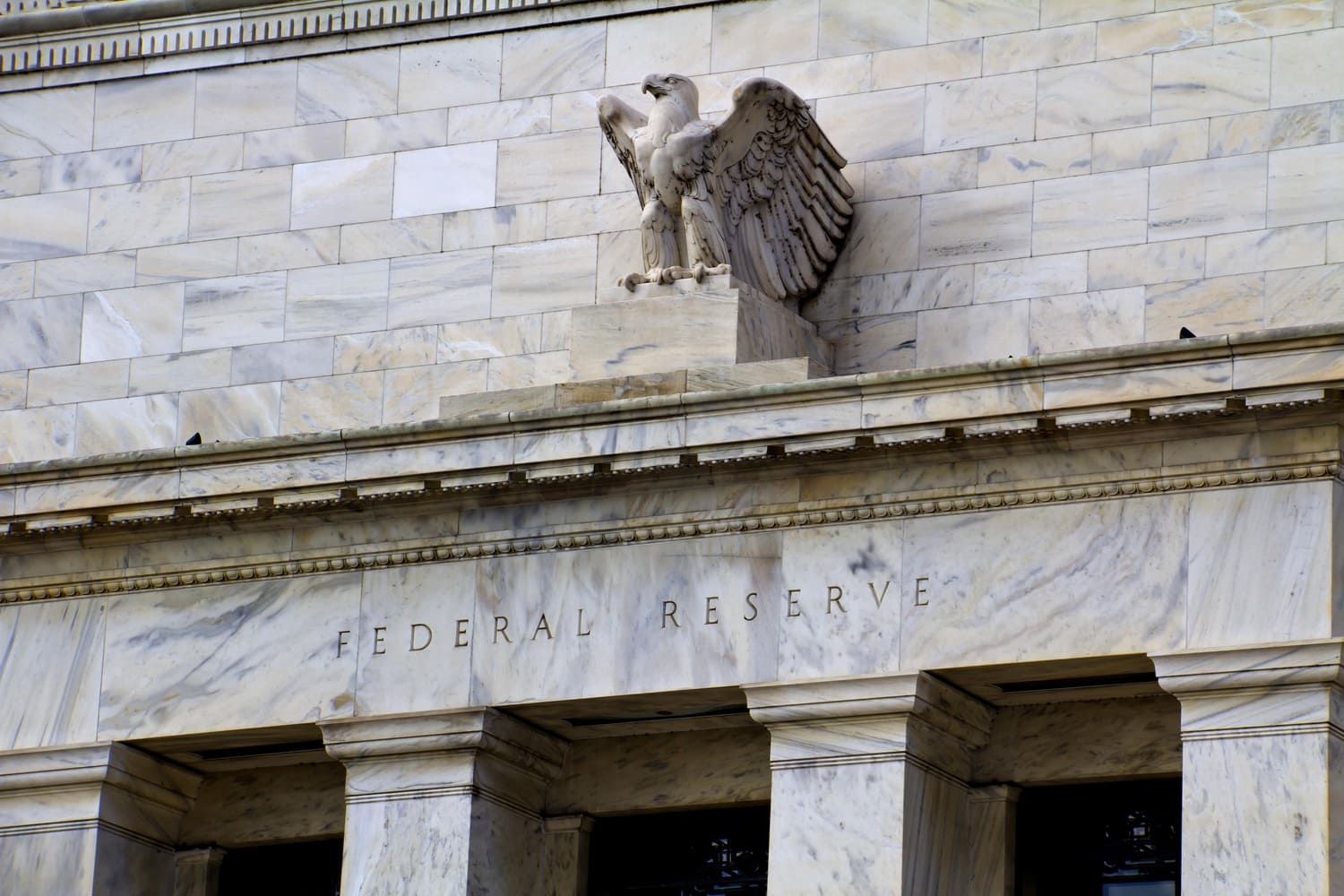 A low-angle shot of the white marble exterior of the Federal Reserve building, featuring the engraved "FEDERAL RESERVE" title and a large stone American eagle sculpture with outstretched wings perched above the entrance.