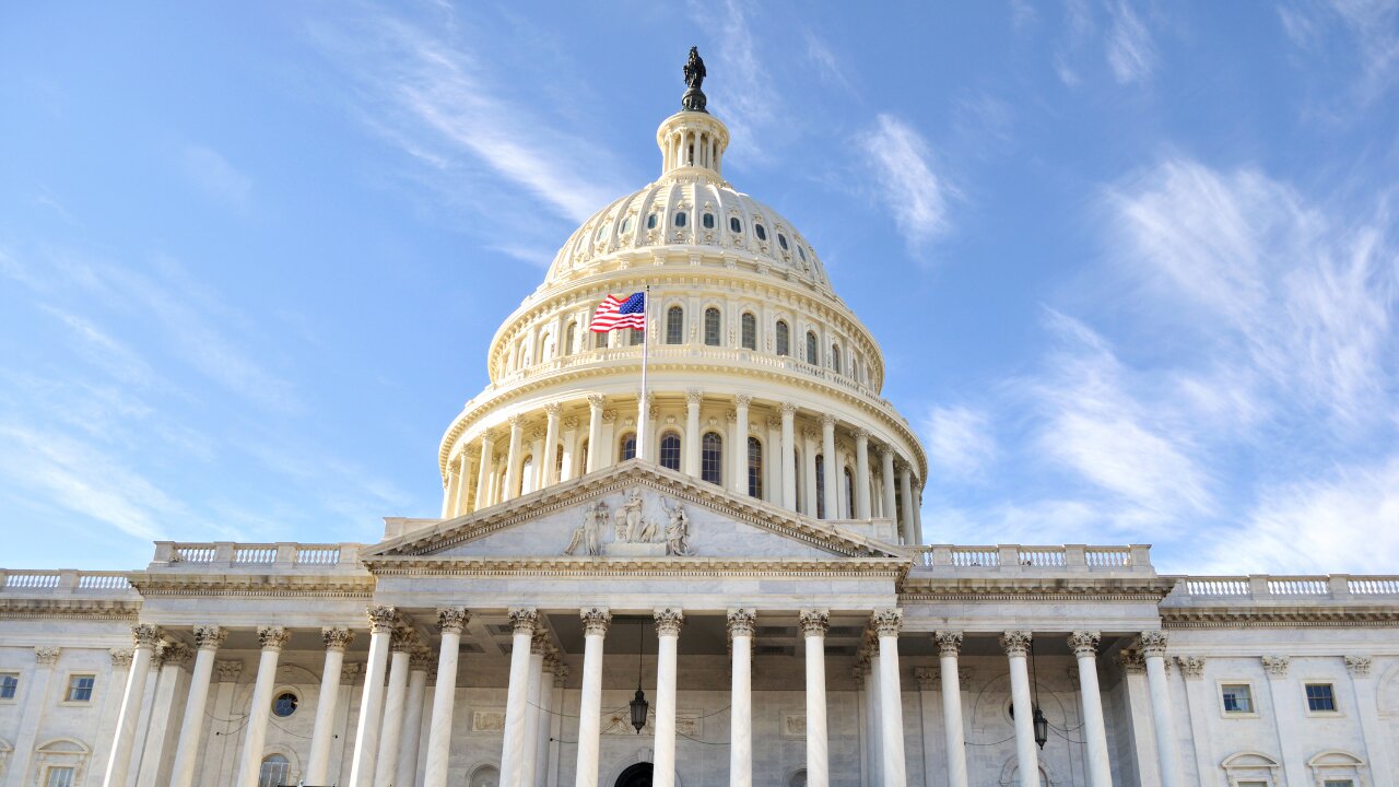 A low-angle shot of the United States Capitol Building in Washington, D.C., featuring the white dome and an American flag waving against a bright blue sky with wispy clouds.
